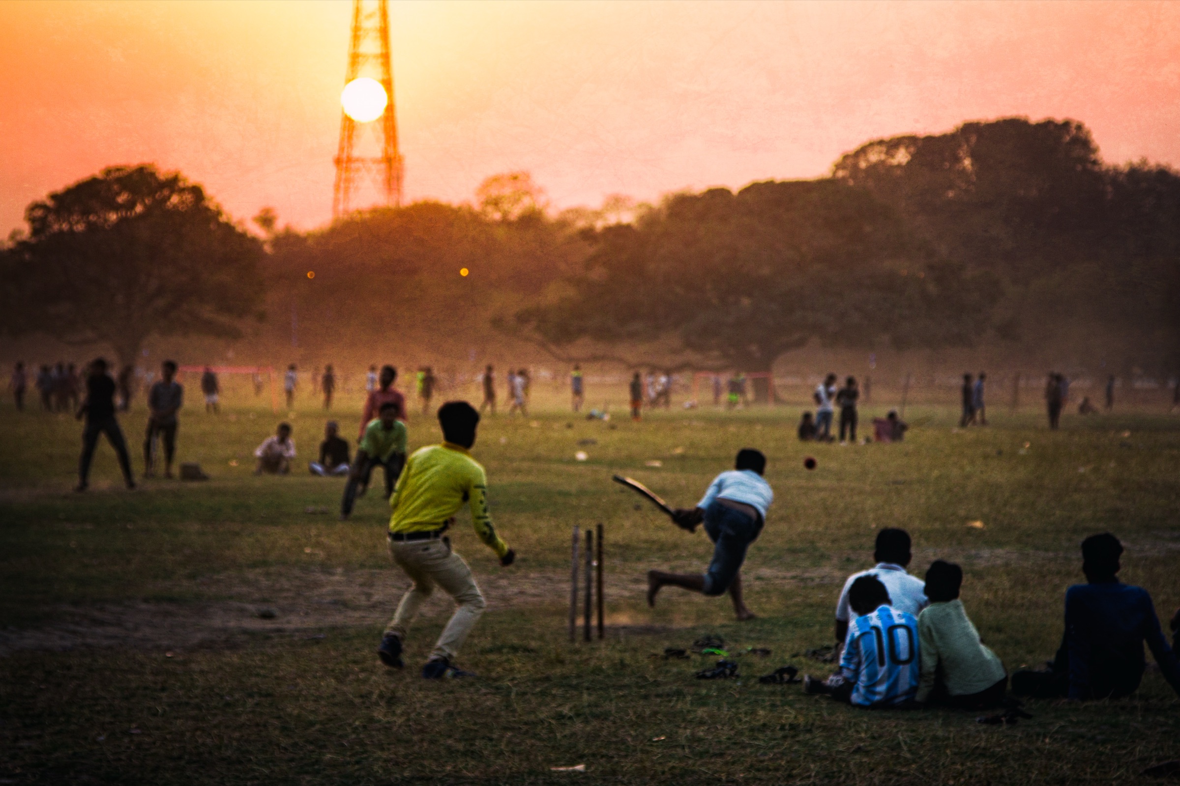 An evening cricket game on the Maidan, the batsman mid-swing as the sun drops behind a lattice radio tower.